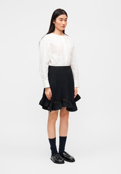 Άσπρο πουκάμισο με κουμπιά, άνετη εφαρμογή και μακριά μανίκια· συνδυασμένο με μαύρη, πλισέ φούστα με διακοσμητικό βολάν και διαφανείς λεπτομέρειες.