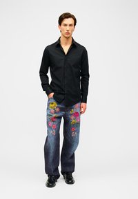 Mann steht, trägt ein schwarzes Langarmshirt und blaue Jeans mit bunter Blumenstickerei, kombiniert mit schwarzen Schuhen.