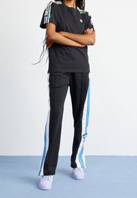 Svart t-shirt med blommig broderi och tre vita ränder på ärmarna, tillsammans med svarta byxor med blå och vita sidostriper.