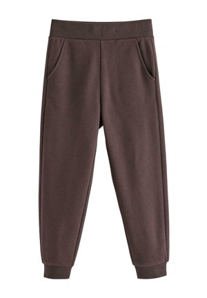 Unge barn iført brun hue, denimjakke over blå langærmet skjorte, brune bukser og røde og hvide sneakers, stående mod hvid baggrund.