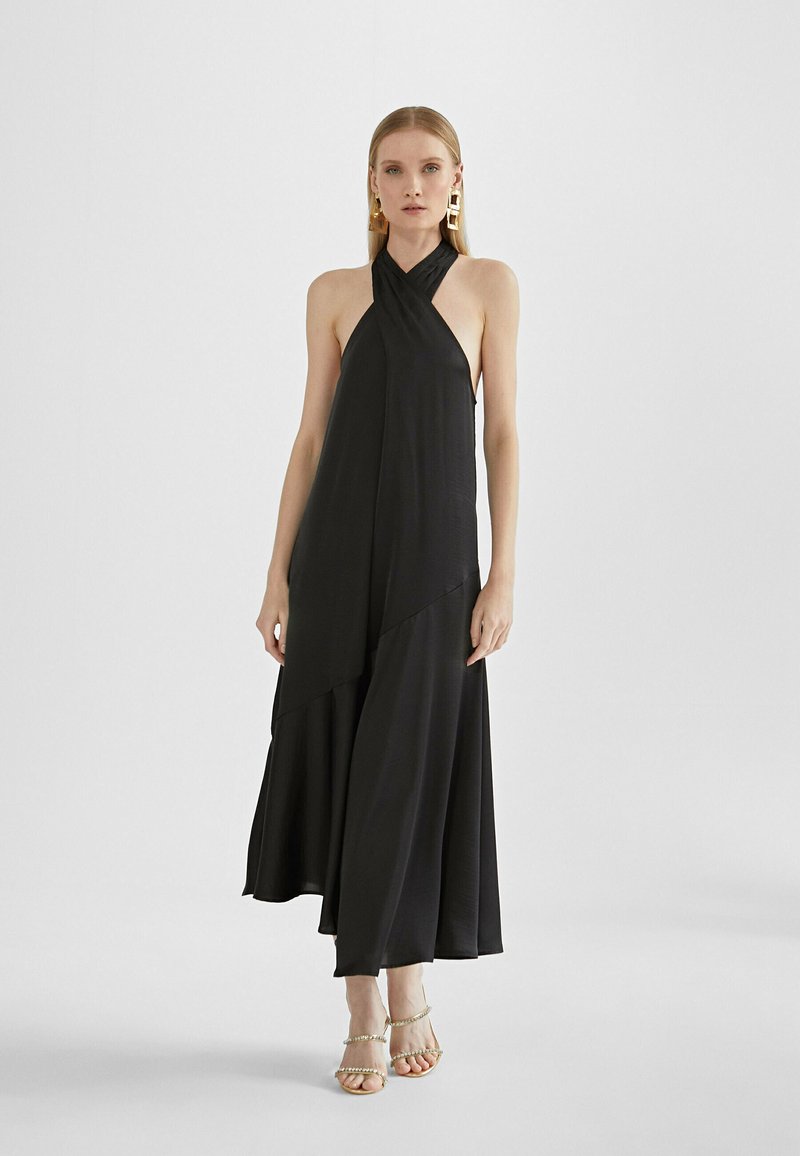 Black halter dress featuring a flowing silhouette, layered design, soft fabric, and a floor-length hem. Complemented by strappy gold sandals.