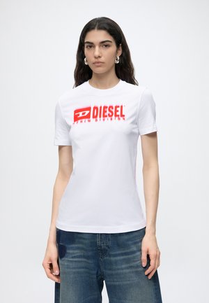 Młoda kobieta z długimi ciemnymi włosami, ubrana w białą koszulkę z logo Diesel i niebieskie dżinsy, stoi na tle jednolitego jasnego tła.