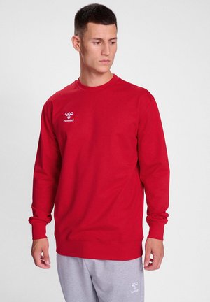 Felpa rossa a maniche lunghe in cotone misto, con scollo rotondo e logo ricamato sul lato sinistro del petto. Il modello indossa pantaloni grigi.