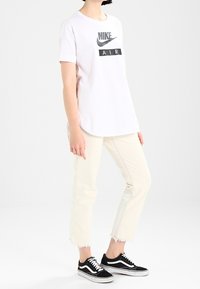 Weißes T-Shirt mit einem grauen Nike-Logo und "AIR" in schwarzer Schrift, kombiniert mit hellbeigen, verkürzten Jeans und schwarzen Vans-Sneakern.