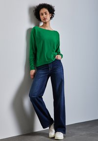 Grünes Langarmshirt mit Boot-Ausschnitt, kombiniert mit dunkelblauen weiten Jeans und weißen Sneakers. Glattes Material mit leichtem Glanz.