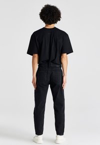 Camiseta negra de algodón y jeans de denim oscuros, de corte recto. La vista trasera muestra las costuras de los hombros y dos bolsillos traseros en los jeans. El calzado son zapatillas blancas.