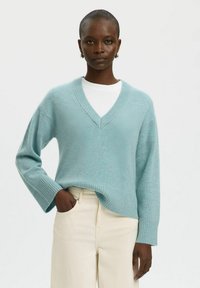 Light teal sweater with a v-neck, ribbed cuffs, and hem. Worn over a white shirt, paired with beige trousers. Soft texture, relaxed fit.