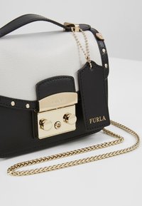Borsa a mano in pelle bianca e nera con ferrature dorate, dotata di chiusura a fermaglio, tracolla a catena e targhetta per bagagli con la scritta "Furla" in rilievo.