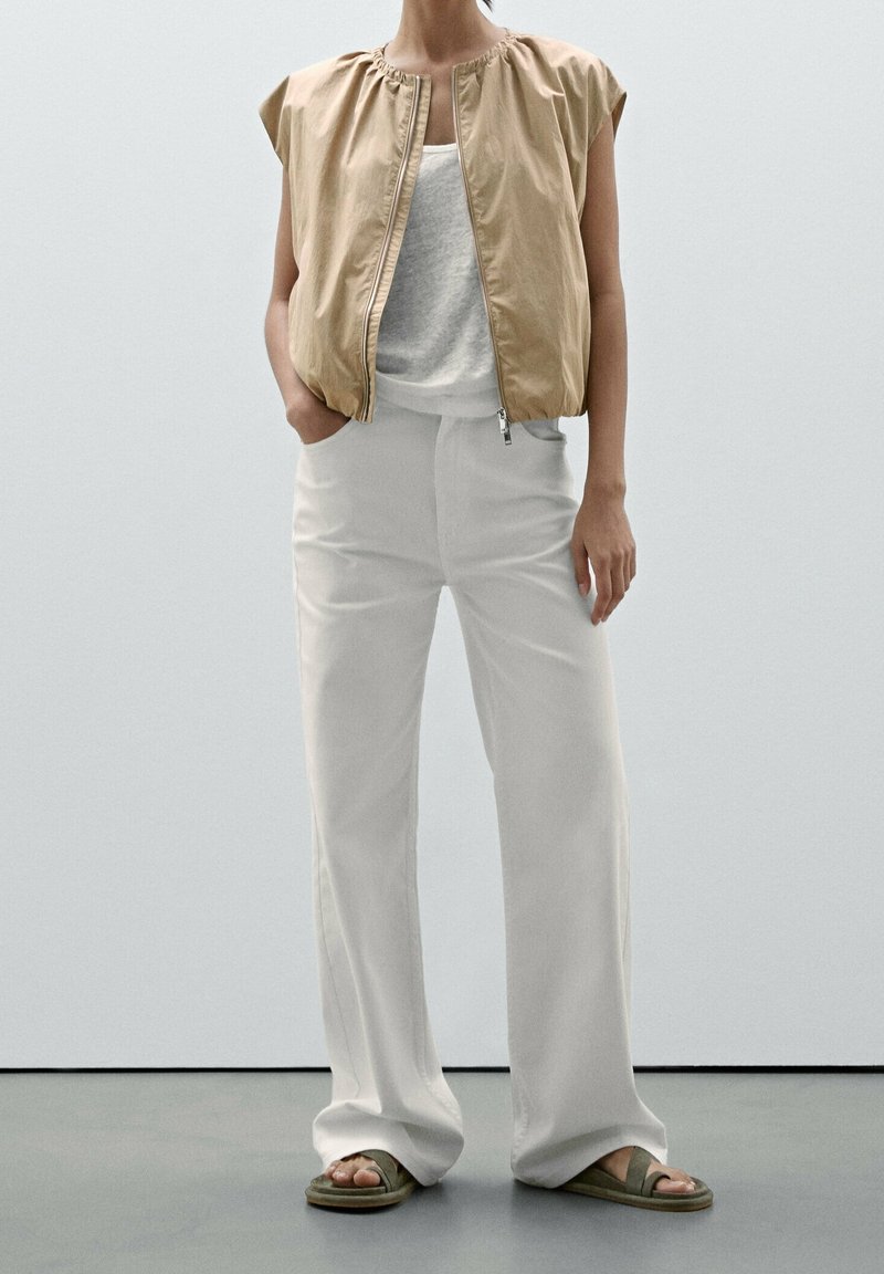 Personne portant une veste zippée beige sans manches sur un haut blanc, un pantalon blanc à jambes larges et des sandales vertes, debout devant un fond clair uni.