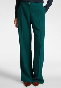 Pantaloni a gamba larga in teal scuro, con una texture liscia, pettorina frontale e chiusura con bottone dorato. Indossati con tacchi a punta marroni.