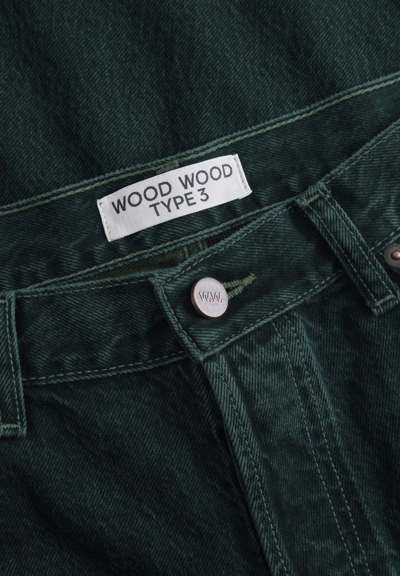 Dark green denim pants featuring a white label with "WOOD WOOD TYPE 3," a metal button, and reinforced stitching along the seams.