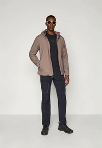 Vyras, vilkintis smėlio spalvos švarką, juodą marškinėlius, juodas kelnes, juodus batus ir baltus akinius nuo saulės, stovi su viena ranka kišenėje.