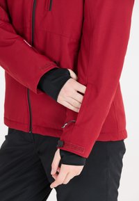 Röd jacka med texturerad yta, dragkedja fram, justerbara handledsmuddar med kardborre, och en figursydd design som är anpassad för rörelse.