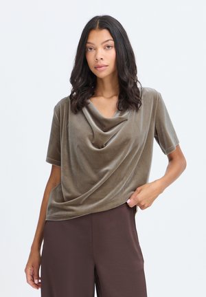 Jeune femme aux cheveux bouclés et foncés portant un haut ample en velours marron avec un col drapé et un pantalon marron foncé sur un fond uni.