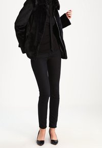 Donna che indossa una giacca nera in pelliccia sintetica, una camicia nera, pantaloni neri slim e tacchi a punta neri, in piedi contro uno sfondo bianco.