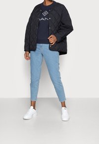 Chaqueta negra acolchada con cierre de botones, sudadera azul marino con logo, jeans claros ajustados y zapatillas blancas, exhibidas contra un fondo liso.