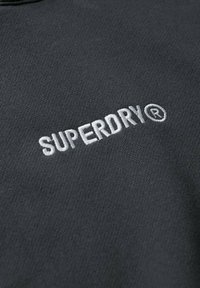 Sudadera gris con el logo bordado "SUPERDRY®" en blanco. La tela tiene una textura suave con un diseño simple y minimalista.