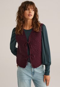 Gilet bordeaux trapuntato con motivo floreale, con legature frontali e scollatura rotonda, indossato sopra una maglietta a maniche lunghe color teal, abbinato a jeans chiari.