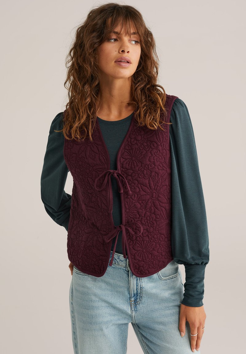 Gilet bordeaux trapuntato con motivo floreale, con legature frontali e scollatura rotonda, indossato sopra una maglietta a maniche lunghe color teal, abbinato a jeans chiari.