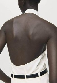Vista trasera de una persona que lleva una blusa sin mangas blanca con la espalda descubierta, combinada con pantalones de cintura negra y un cinturón negro.