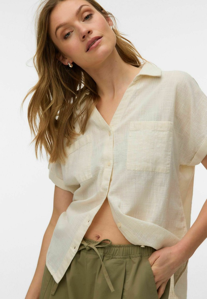 Femme portant une chemise beige clair à manches courtes et boutonnée, ample, avec un pantalon à cordon de serrage vert olive, main dans la poche, tête inclinée, fond clair.