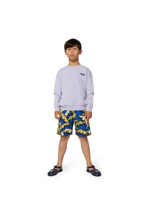 Sweatshirt lilas à col rond avec des épaules tombantes, associé à un short multicolore avec un motif de feuilles tropicales. Des sandales sombres complètent le look.
