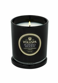 En svart ljusbeh�ållare med etiketten "Voluspa Burning Woods", som har en gyllene cirkulär design och vit vax, står på en matchande bas med en tänd väge.