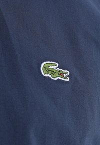 Marineblauwe stof met een geborduurd groen krokodillenlogo, geaccentueerd met rode details aan de zijkant. De textuur is glad en licht getwilld.