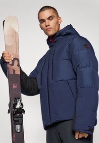 Navy blue puffer jacket with a hood, featuring black accents, zippered pockets, and a smooth texture, paired with skis and bindings.