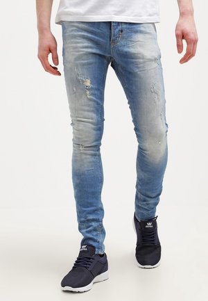 Personne portant un jean skinny déchiré bleu clair et des baskets Supra noires marchant sur un fond blanc uni.