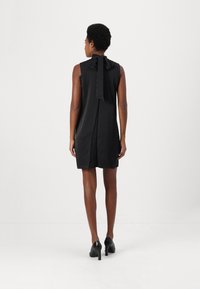 Svart ärmlös klänning med en rosett på baksidan, slät textur, knäskuren längd och kompletterad med svarta högklackade skor.