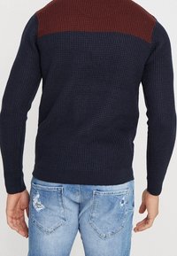 Mörkblå stickad tröja med en strukturerad design och vinröda axeldetaljer, tillsammans med ljusblå slitna jeans som visar fickdetaljer.