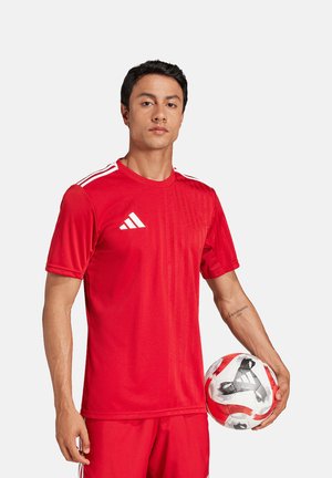 Mann in roter Sportkleidung hält einen weiß-roten Fußball vor einem einfarbigen Hintergrund.