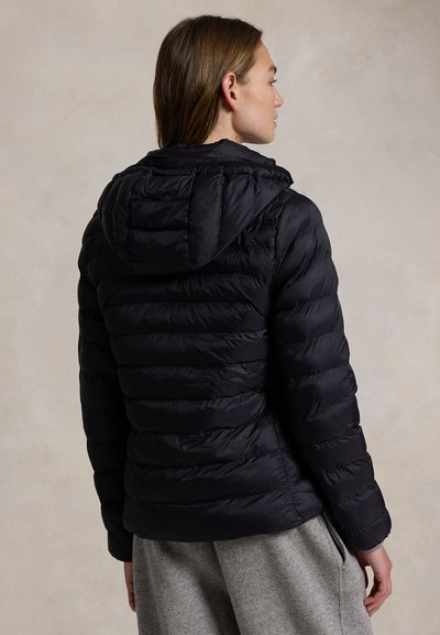 Osoba oblečená v černé prošívané bundě s kapucí a šedých teplácích, čelem od fotoaparátu na neutrálním pozadí.