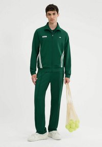 Zielony dres z zamkami, pasek boczny i pasujące spodnie. Zawiera siateczkową torbę z żółtymi piłkami tenisowymi. Białe obuwie.