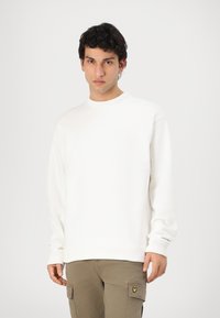 Hvit sweatshirt med ribbestrikket rund hals og mansjetter, med en liten logo detalj. Kombinert med lysebrune cargobukser. Glatt tekstur.