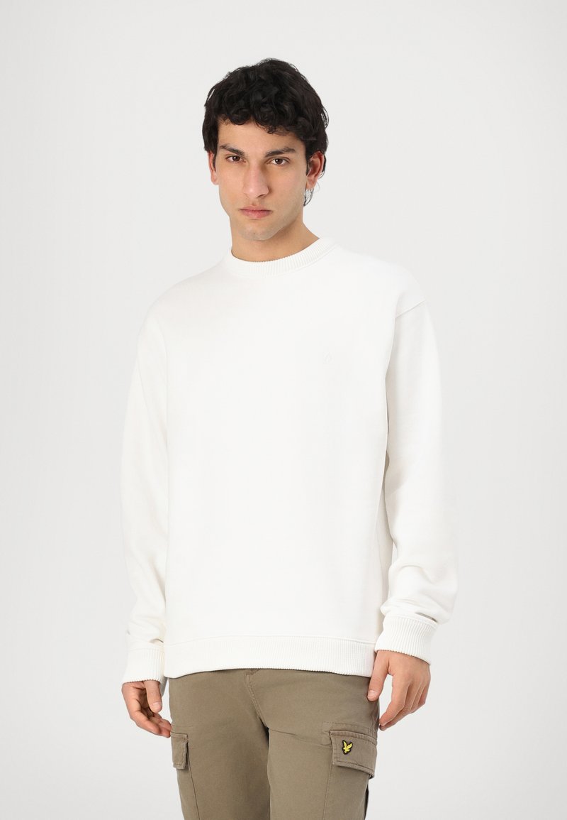 Hvit sweatshirt med ribbestrikket rund hals og mansjetter, med en liten logo detalj. Kombinert med lysebrune cargobukser. Glatt tekstur.