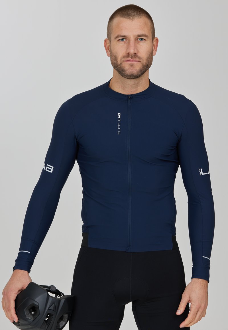 Navyfarbendes Radtrikot mit langen Ärmeln, Frontreißverschluss und Markenlogo-Akzenten. Getragen mit schwarzen Fahrradhosen und einem schwarzen Helm.