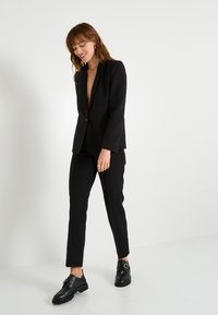 Blazer noir avec un décolleté en V profond et une coupe ajustée, associé à un pantalon noir slim. Le modèle porte des chaussures noires, sur un fond blanc uni.