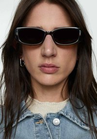 Sorte rektangulære solbriller med tykke stel, båret med en lys blå denimjakke og en struktureret cremet sweater. Ingen synlige mønstre eller accenter.