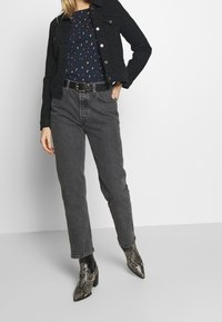 Svart denimjacka som matchas med en marinblå mönstrad skjorta, grå högmidjade jeans och metalliska boots med ormskinna.