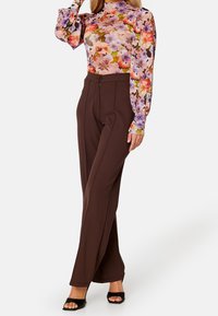 Långärmad blommig blus i rosa, lila och gröna nyanser med puffärmar, matchad med högmidjade bruna skräddarsydda byxor och svarta sandaler.