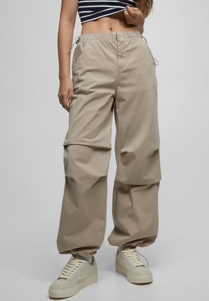 Person wearing beige cargo pants and cream sneakers, holding up a striped navy and white crop top, against plain gray background.