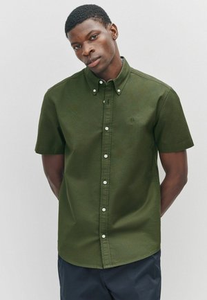 Chemise à manches courtes boutonnée en coton vert olive. Comprend un col classique, des boutons blancs et un ourlet légèrement arrondi. Pas de motifs visibles.