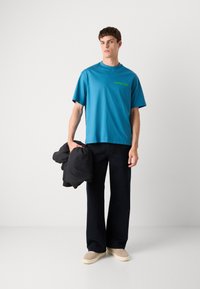 Τυρκουάζ βαμβακερό μπλουζάκι με κοντά μανίκια, πράσινο λογότυπο, σετάρισμα με μαύρο παντελόνι φαρδιάς γραμμής και μια μαύρη μπουφάν που κρατείται στο χέρι.