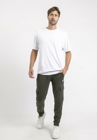 T-shirt blanc à manches courtes associé à un pantalon cargo vert foncé avec de multiples poches, complété par des baskets blanches et un branding minimal.