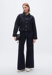 Sötét farmer rövidített dzseki nagy foltzsebekkel, ezüst gombokkal és hegyes gallérral; bő szárú sötét farmernadrággal és egy kis fekete táskával párosítva.