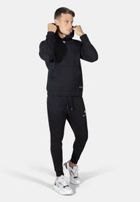 Felpa nera con tasca frontale e cappuccio regolabile, abbinata a pantaloni jogger neri a gamba affusolata. Accenti in bianco sul logo in entrambi i capi.