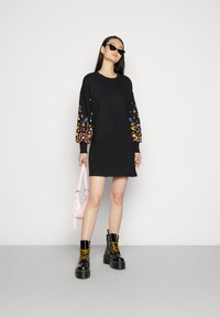 Robe sweat noir avec broderie florale colorée sur les manches longues ; associée à des bottes de combat noires et un sac à bandoulière rose.