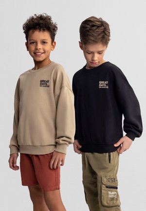 Dwóch chłopców stoi, jeden uśmiecha się do aparatu, ubrany w beżową bluzę i czerwone szorty, drugi patrzy w dół, ubrany w czarną bluzę i zielone spodnie cargo.